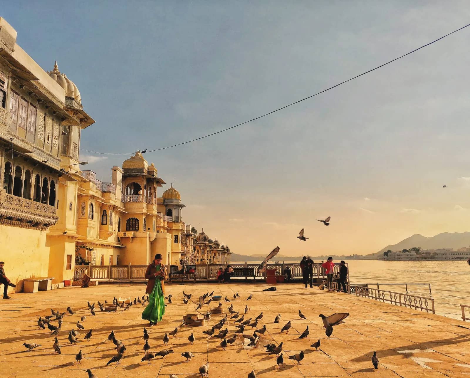 A woman in a green dress walking across a lakeside ghat in Udaipur, with pigeons on the stone