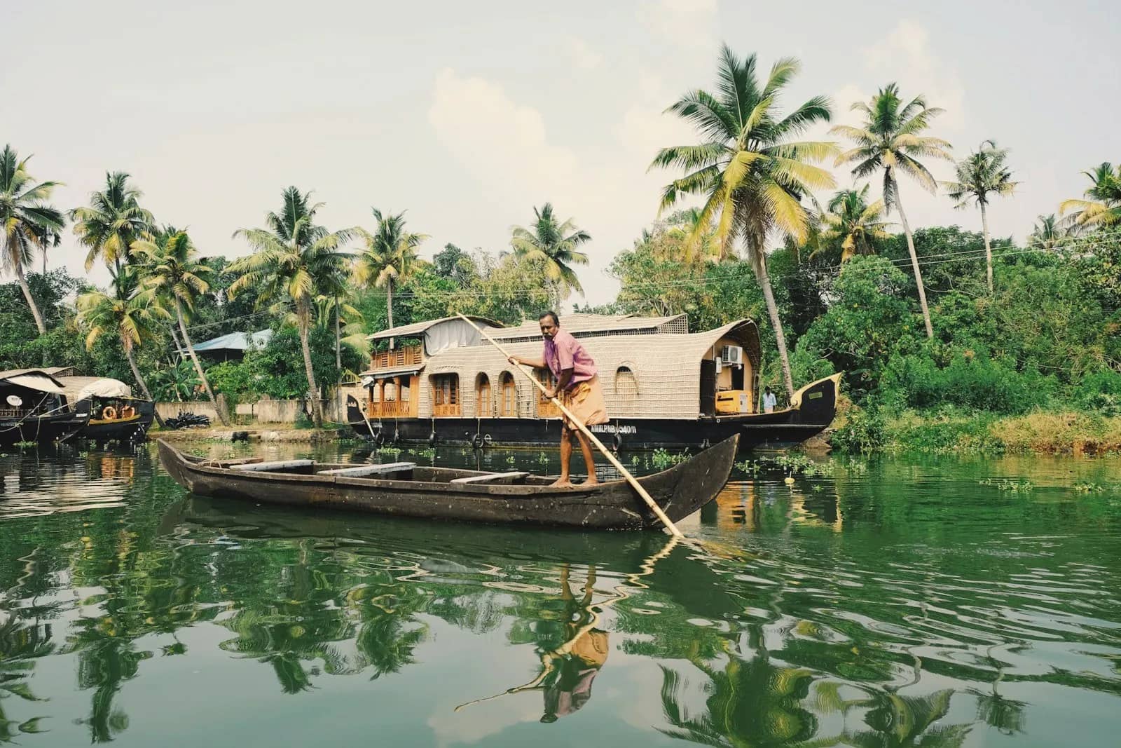 A boatman poling a boat past a houseboat and palm trees on the Kerala backwaters